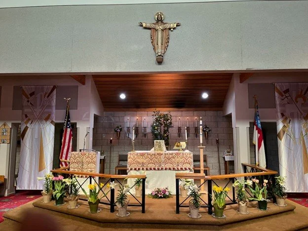 Inside a church altar area decorated for a religious service, with a statue of Jesus on the cross above and American flags on either side, surrounded by potted flowers.