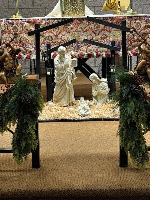 Nativity scene display with statues of Mary and Joseph, baby Jesus, and angels, decorated with greenery and pinecones, set in a church or religious setting.