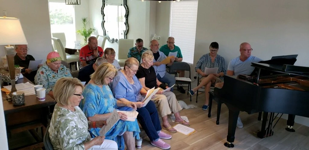A group of people sitting in a room, singing or reading from sheet music, with a pianist playing a grand piano.