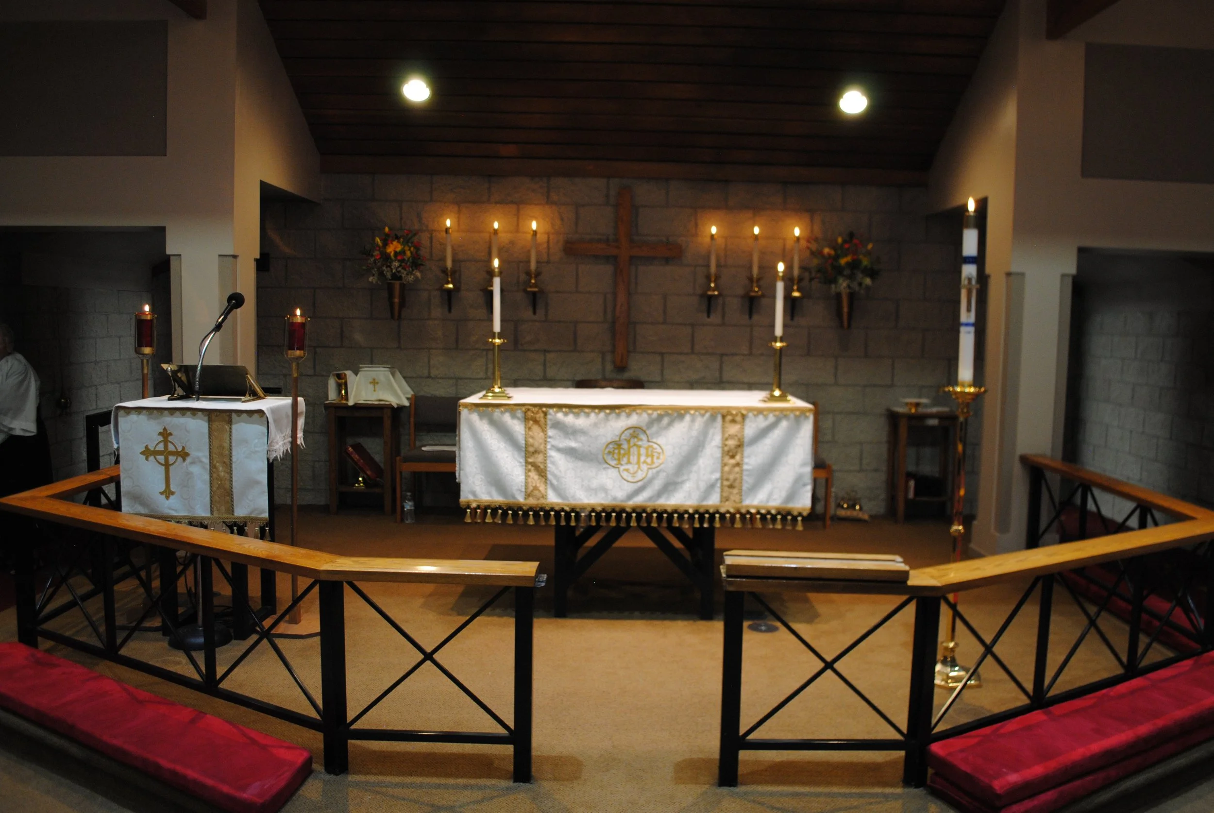 Interior of a small church or chapel with an altar, cross, candles, and floral arrangements, viewed from the pews.