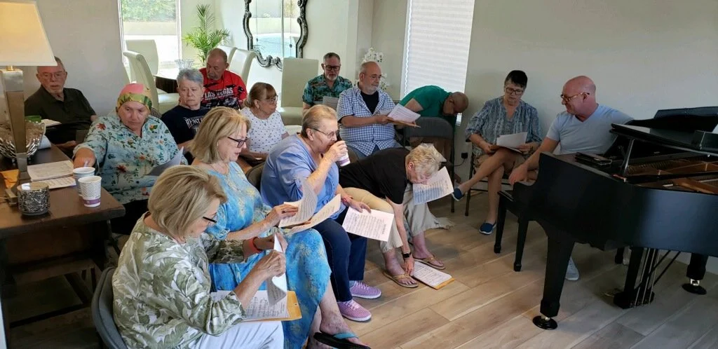 A group of seniors and a few adults gathered in a living room, sitting in chairs and on the floor, holding sheets of paper, with a black grand piano in the corner, and a window with blinds in the background.