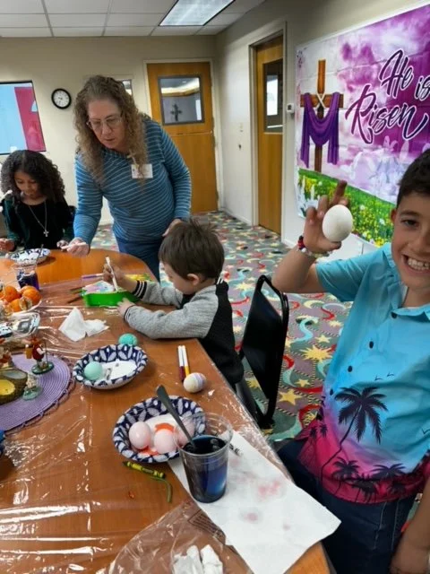 Children and a woman engaging in an arts and crafts activity at a table with decorated eggs, in a colorful room with a cheerful sign that says 'He is Risen' and has a cross on the door.