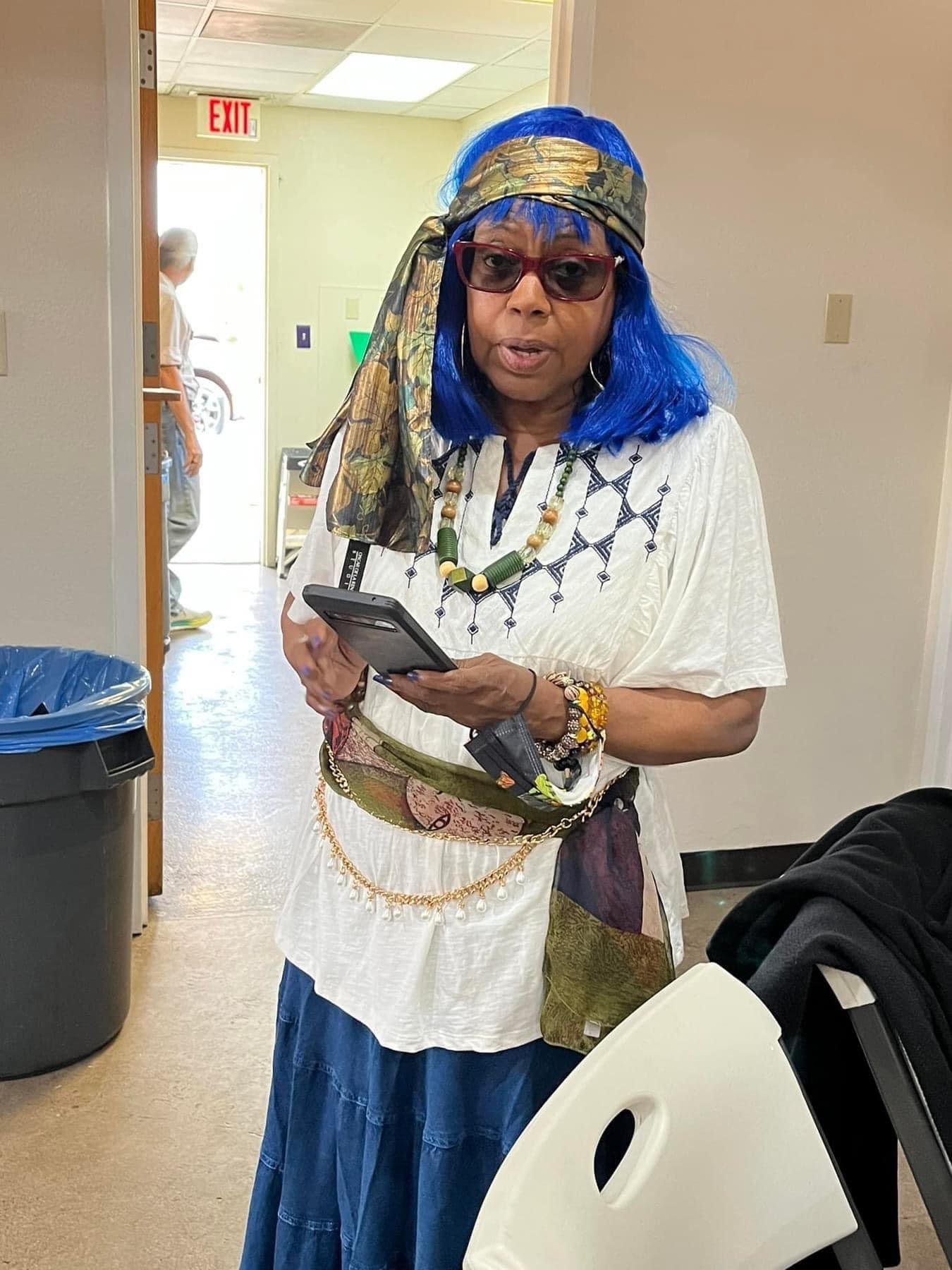 A woman with blue hair, wearing sunglasses, a patterned headscarf, and a white blouse, holding a smartphone, standing indoors near a trash can and a white chair.