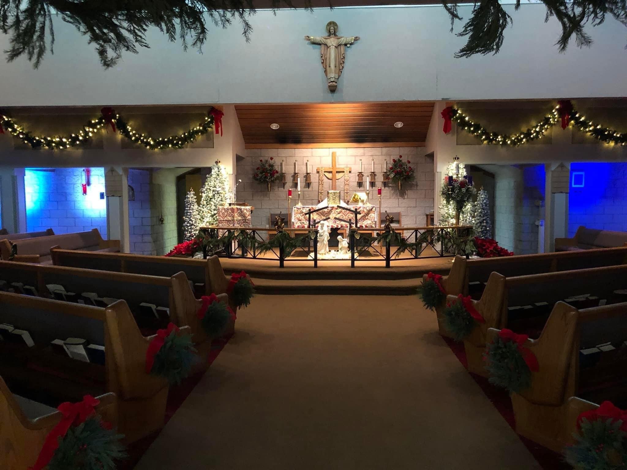 Interior of a church decorated for Christmas with garlands, poinsettias, Christmas trees, and lit candles, with a large wooden cross and religious figures at the altar.