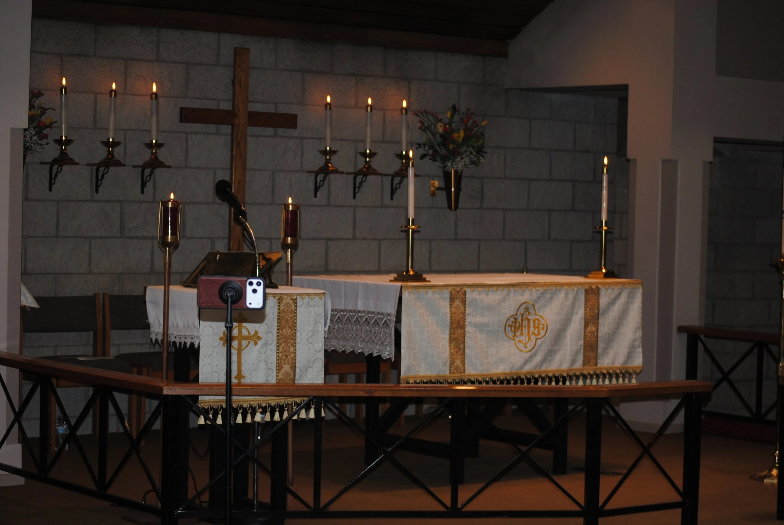 Interior of a church altar setup with lit candles, a wooden cross, flowers, and embroidered cloths.
