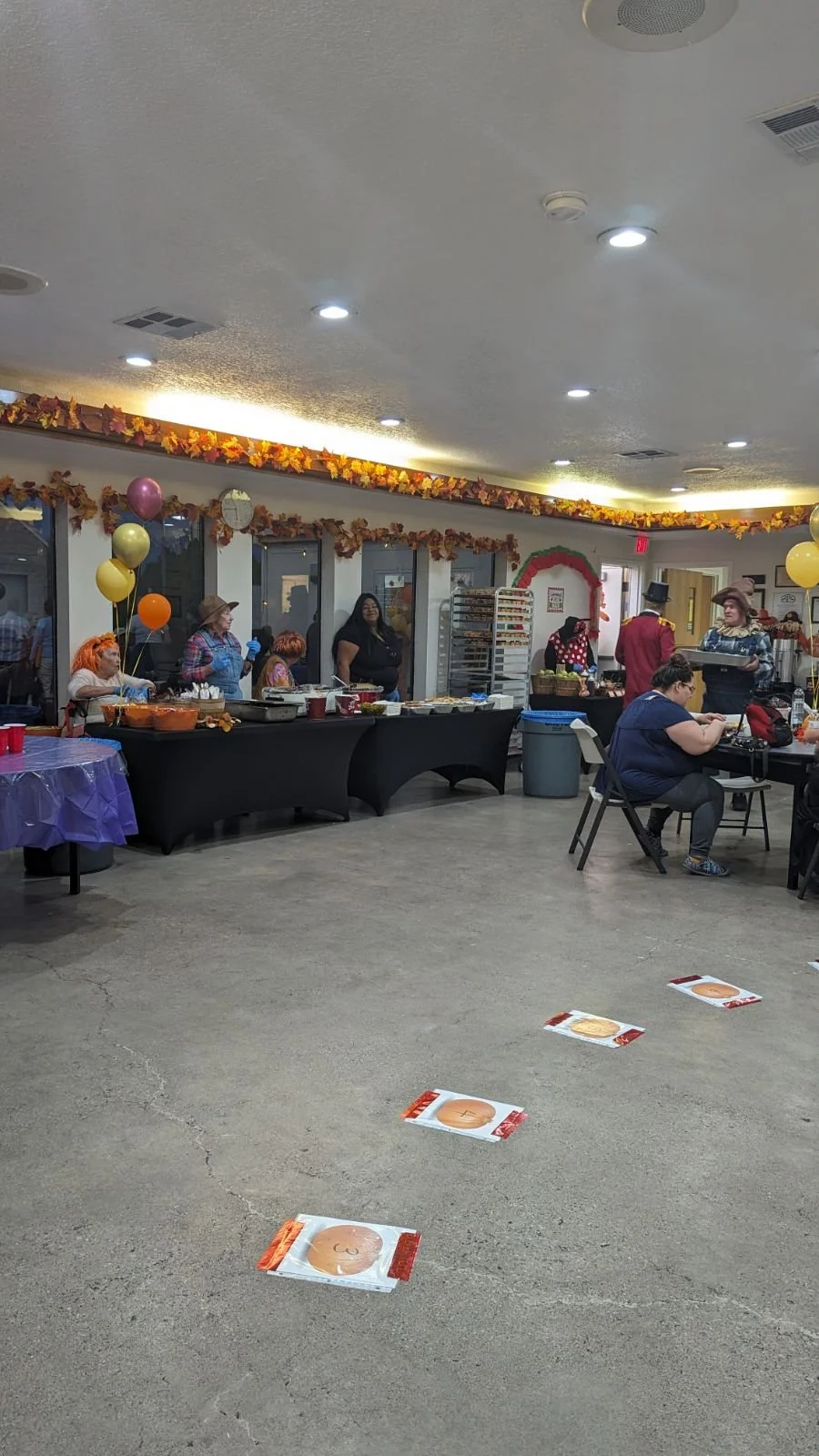 Indoor gathering with Halloween decorations, tables, and people, some dressed in costumes. Floor has four placemats with pumpkin designs.