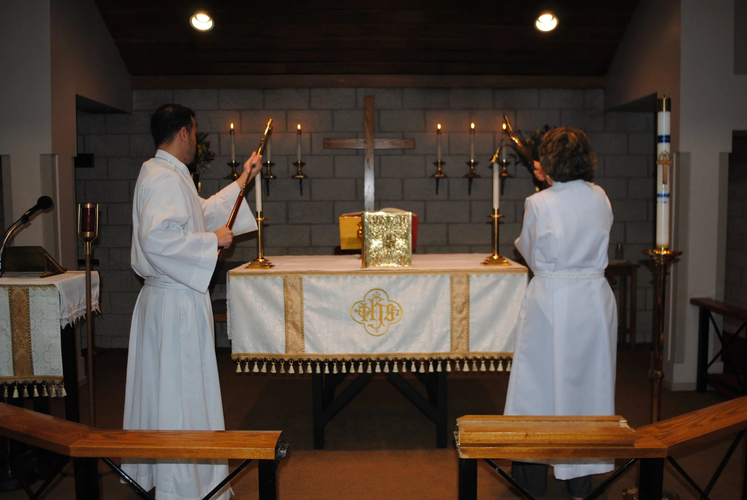 Two priests in white robes conducting a church service at an altar with lit candles, a cross, and religious items inside a church.