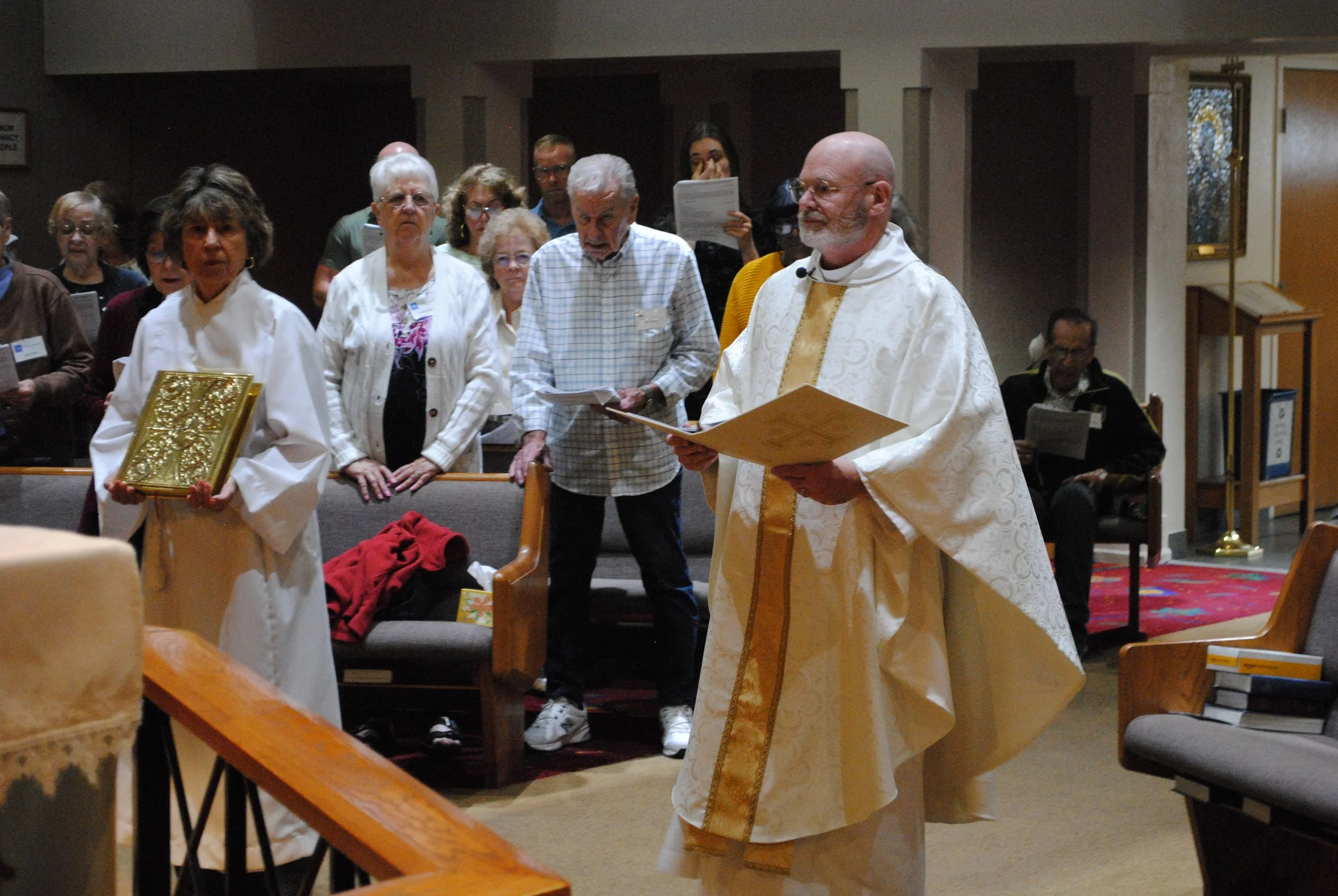 A priest dressed in white and gold robes leading a religious service inside a church with a group of mostly elderly people gathered around him.