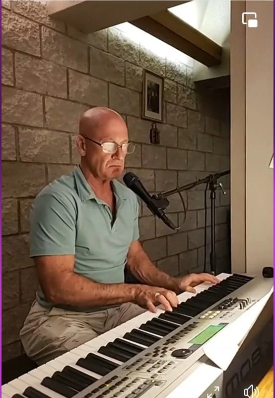An elderly man with glasses and a green polo shirt playing a digital keyboard with a microphone in front of him, against a brick wall.