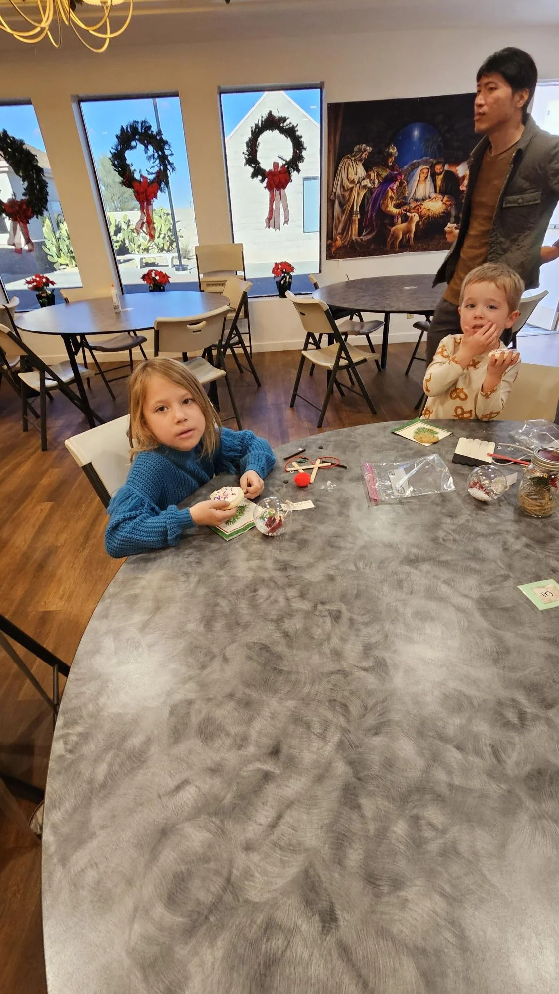 Two young children sitting at a large round table with Christmas cookies and decorations, with a man standing nearby, in a festive room with holiday wreaths and Christmas artwork.
