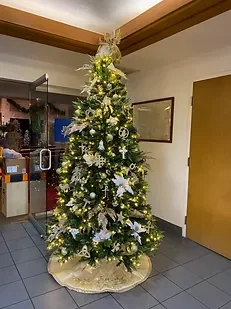 Decorated Christmas tree with white and gold ornaments and lights, placed indoors on a tiled floor.
