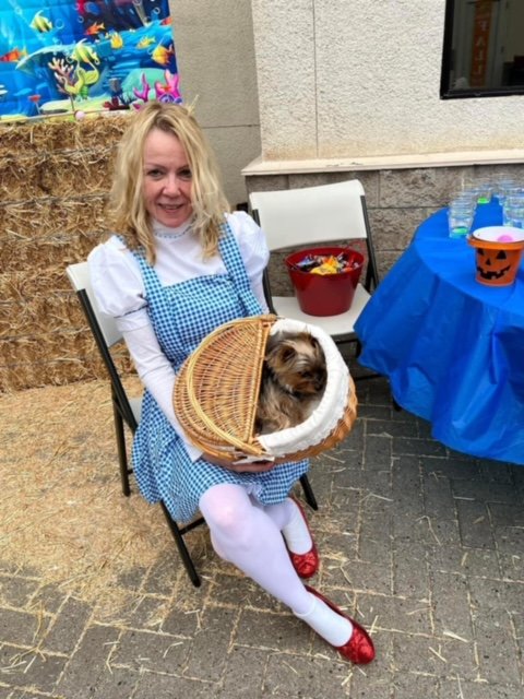 Woman dressed as Dorothy from The Wizard of Oz holding a small dog in a basket, sitting near a table with a blue tablecloth and colorful cups, at a festive outdoor event.