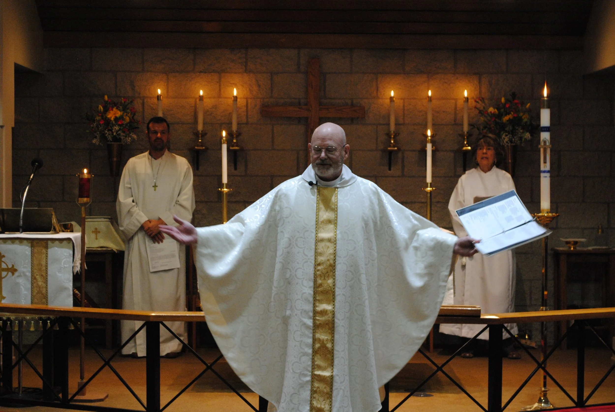 A priest in white religious robes with gold accents leading a church service, standing with arms outstretched in front of an altar decorated with candles, flowers, and a wooden cross, with two assisting clergy members in the background.