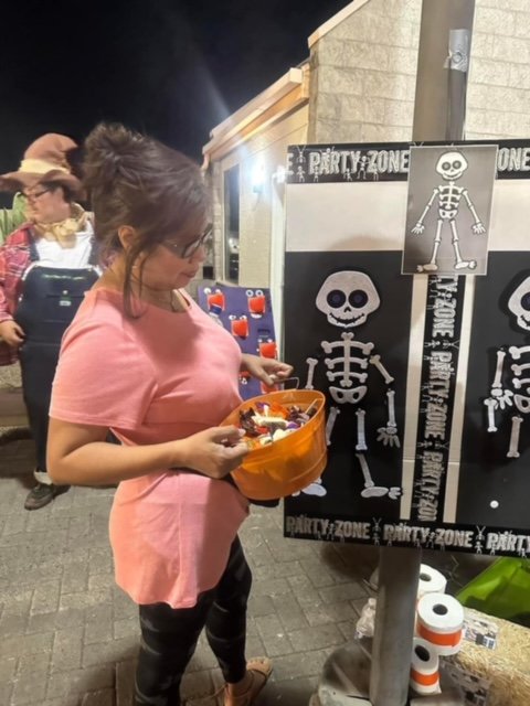 A woman wearing a pink shirt and glasses is collecting candy from a Halloween display with skeleton images and the words 'Party Zone.' The display is outdoors at night, and other people are visible in the background.