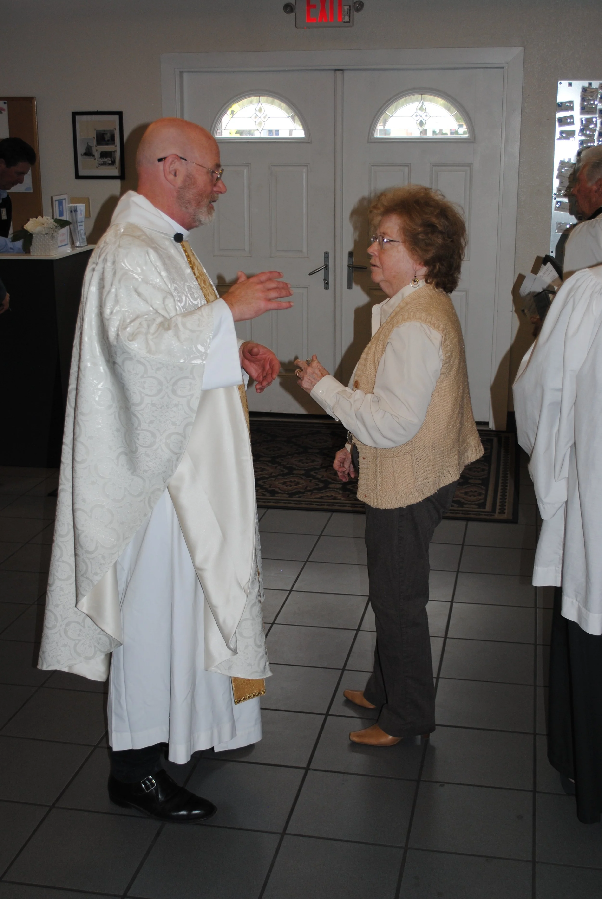 A priest and an elderly woman are having a conversation inside a building near the entrance door. The priest is dressed in white religious robes, and the woman is wearing a beige vest over a white blouse. Other people are present in the background, a