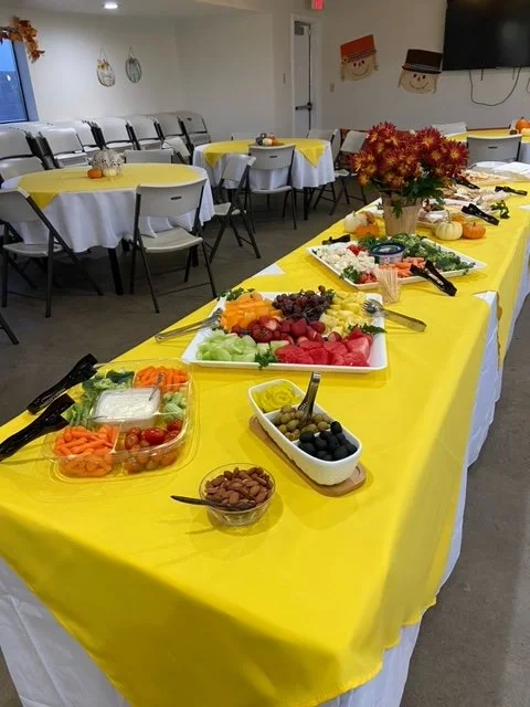 Buffet table with yellow tablecloth displaying a vegetable platter, fruit, olives, and nuts, with a vase of red and orange flowers in the center, set in a decorated event space with yellow and white tables and chairs.