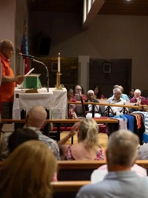 A woman speaking at a church service with elderly attendees seated, some wearing patriotic colors, in a church with American and Christian symbols.