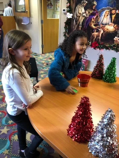 Two young girls are sitting at a table decorated with miniature Christmas trees, with a Nativity scene picture on the wall behind them.