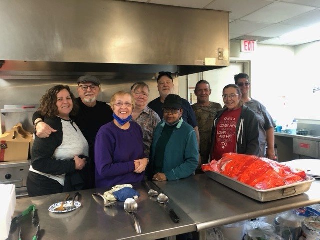 Group of eight people standing in a kitchen, smiling, with a large tray of raw fish covered in red plastic wrap on a stainless steel counter.