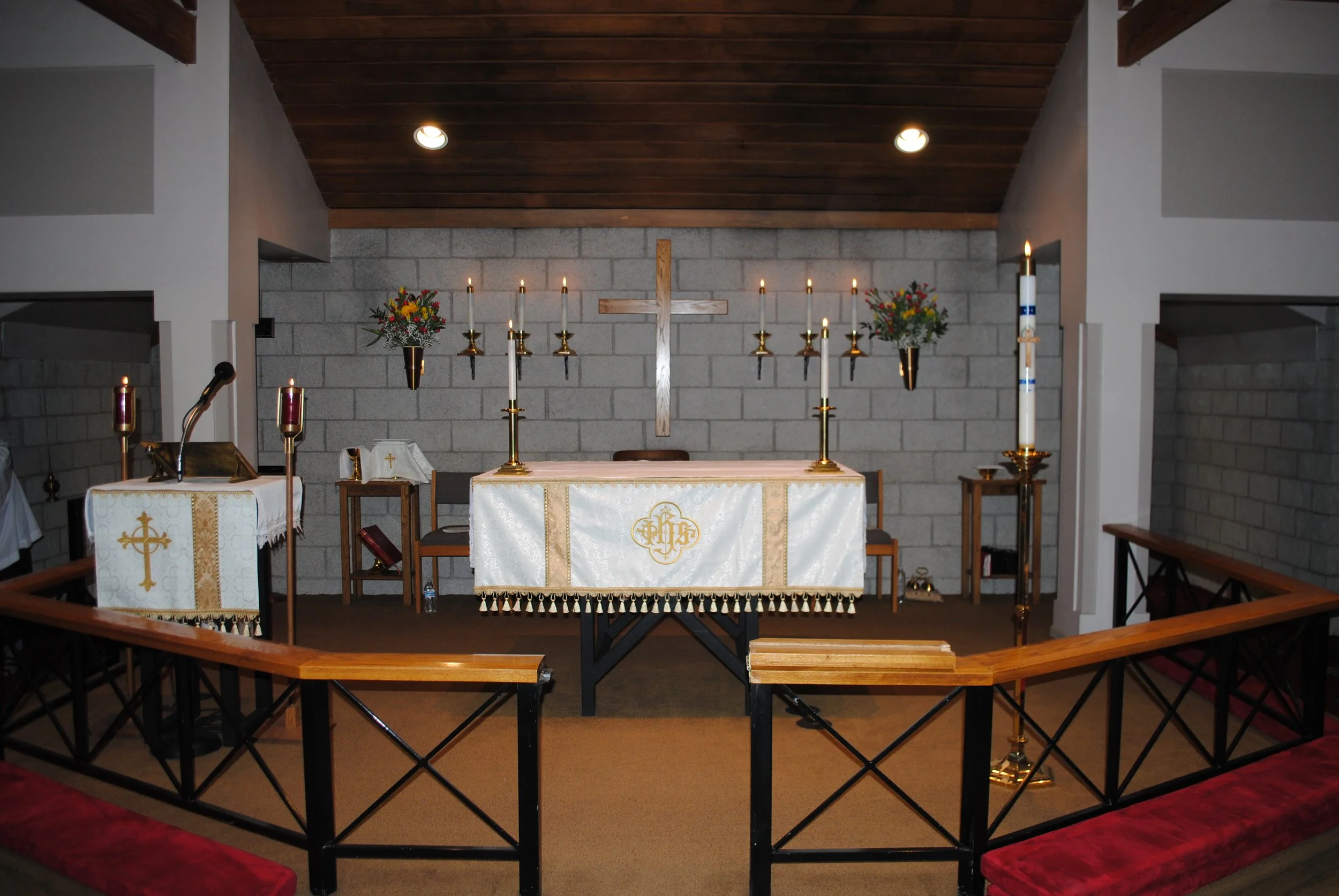 An altar in a church with a wooden cross, candles, floral arrangements, and religious items, surrounded by a wooden railing.
