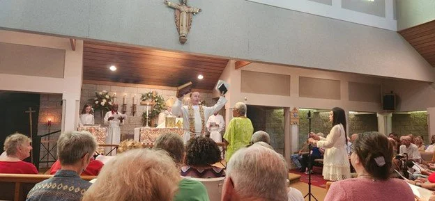 A priest and a woman standing in front of a congregation during a church service. The priest is raising his arms, and there is a cross and altar in the background. People are seated, watching the service.