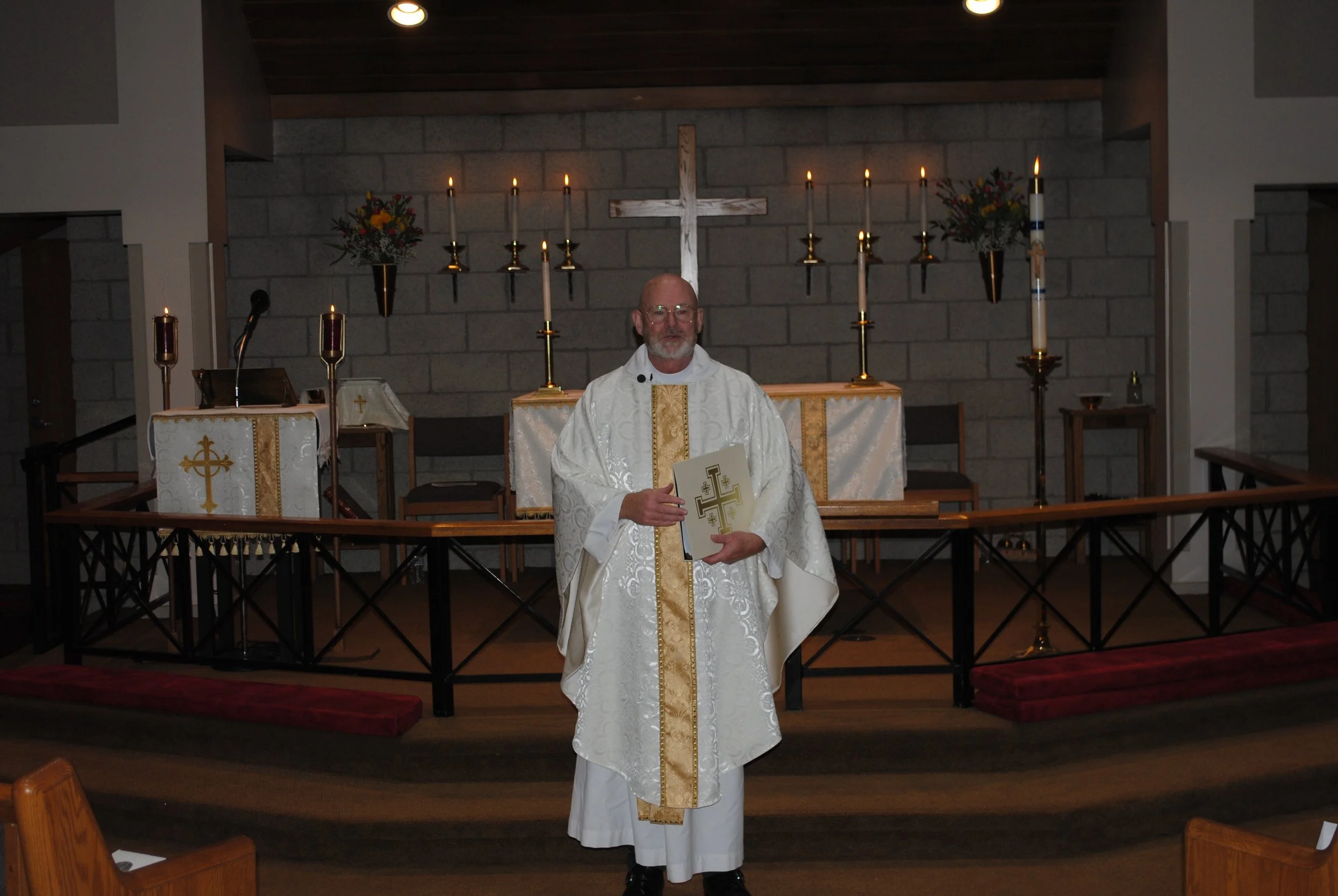 A priest standing in front of altar inside a church, holding a book with religious symbols, dressed in white vestments with gold accents.