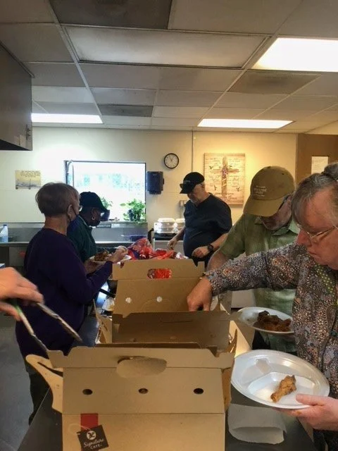 A group of people at a food distribution or cafeteria, lined up to receive meals from boxes on a counter.