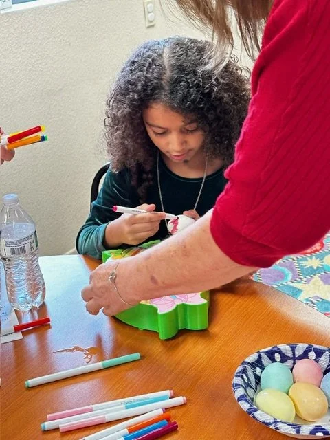 Child with curly hair receiving a paintbrush from an adult at a table with colored markers, a water bottle, and pastel-colored eggs.