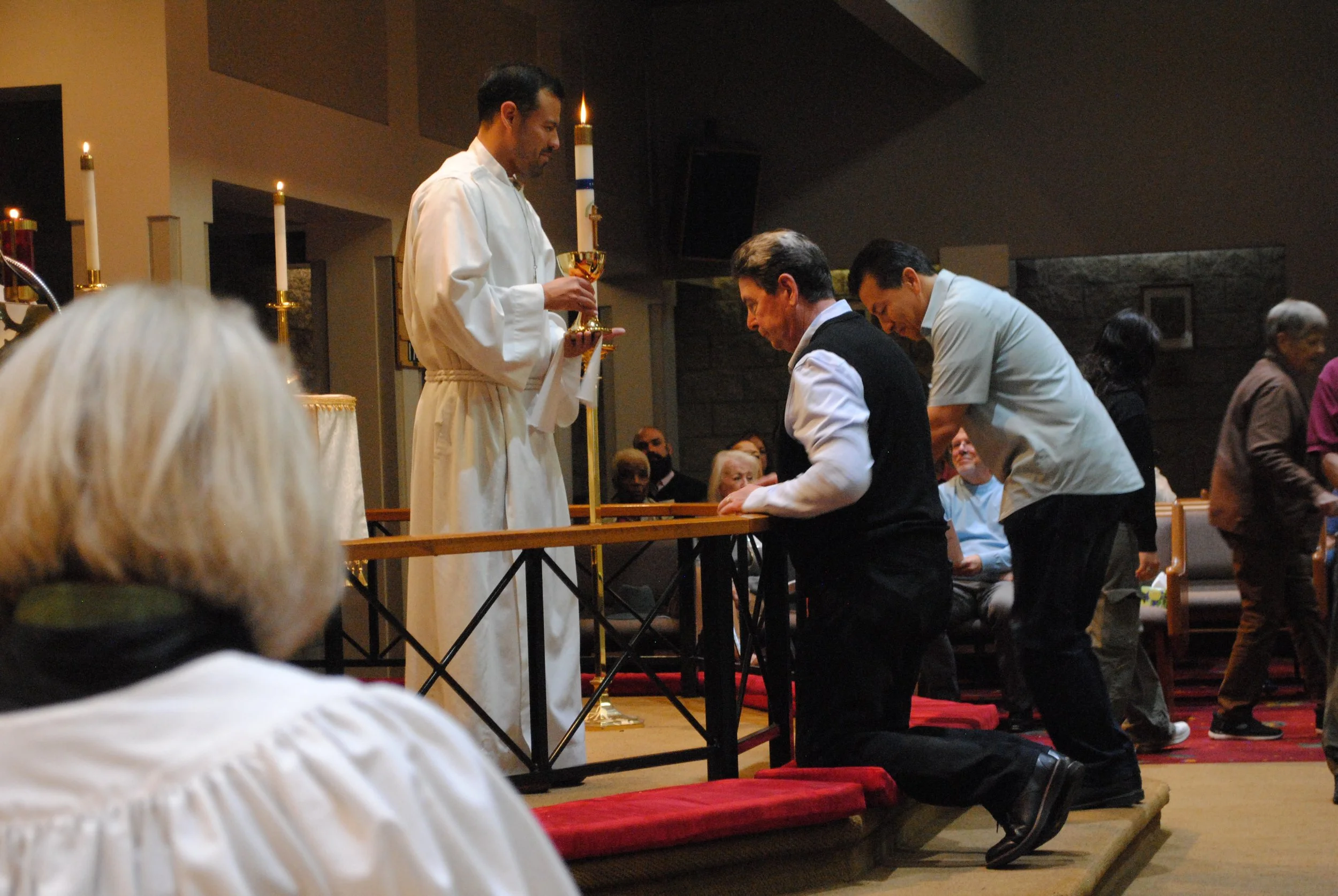 A priest or clergy member in white robes holding a chalice appears to be participating in a religious ceremony. Several individuals are kneeling before the altar, many with their heads bowed or kneeling on red cushions, inside a church or chapel. Oth