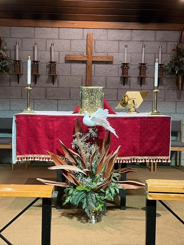 Inside a church, an altar decorated with a red tablecloth, a white dove sculpture, candles, a cross on the brick wall, and floral arrangements.