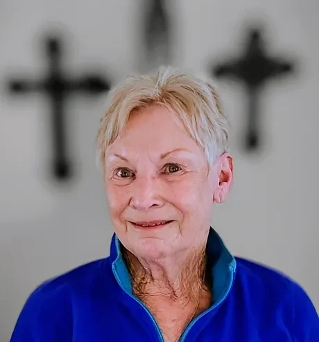A smiling elderly woman with short blonde hair, wearing a blue zip-up jacket, standing in front of a blurred background with crosses.