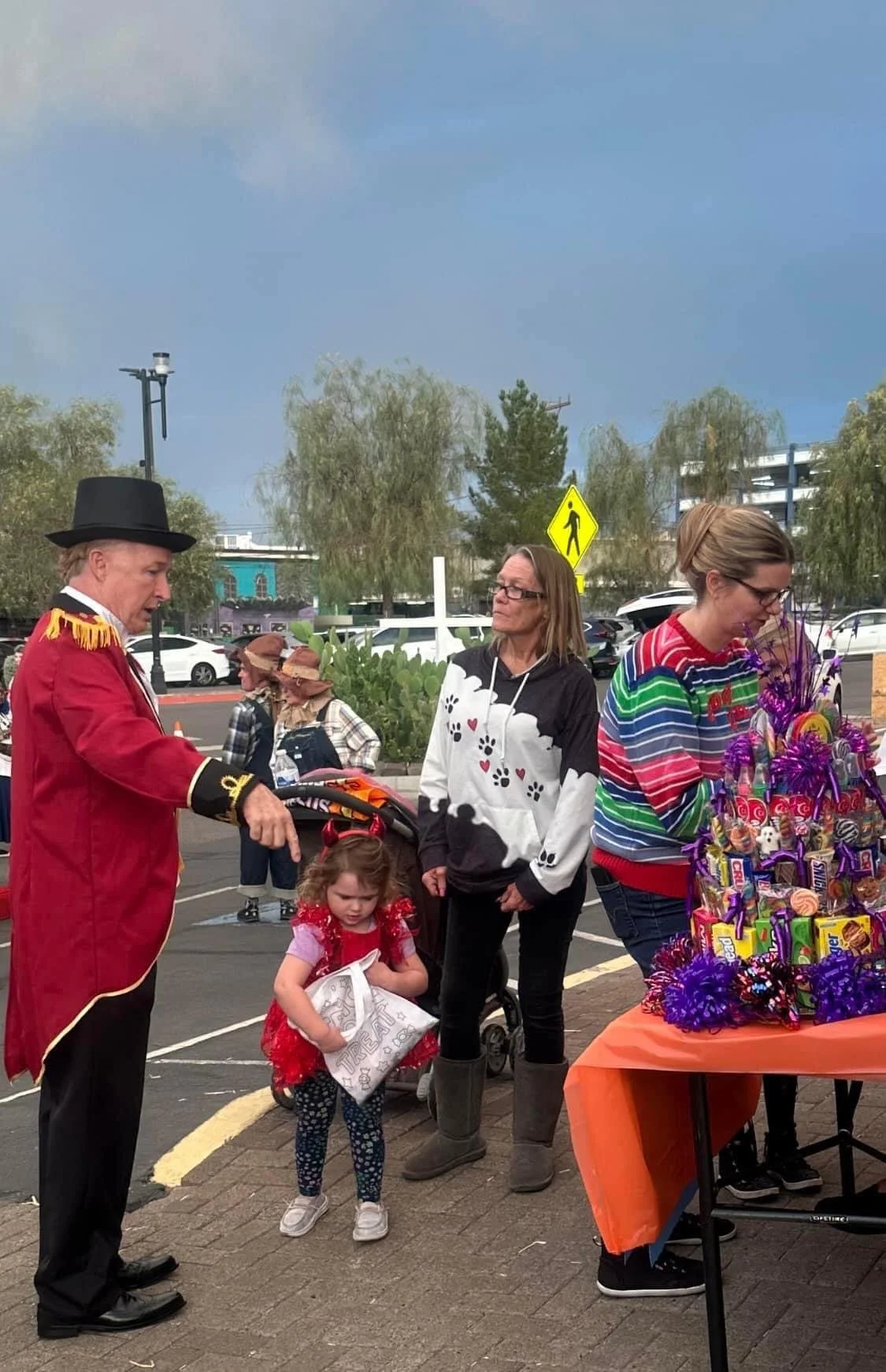A man dressed as a circus ringmaster, a woman, and a young girl at an outdoor event with a table full of candy and Easter eggs. The girl is holding a white bag, and the woman wears a colorful striped sweater. In the background, there are trees, parke