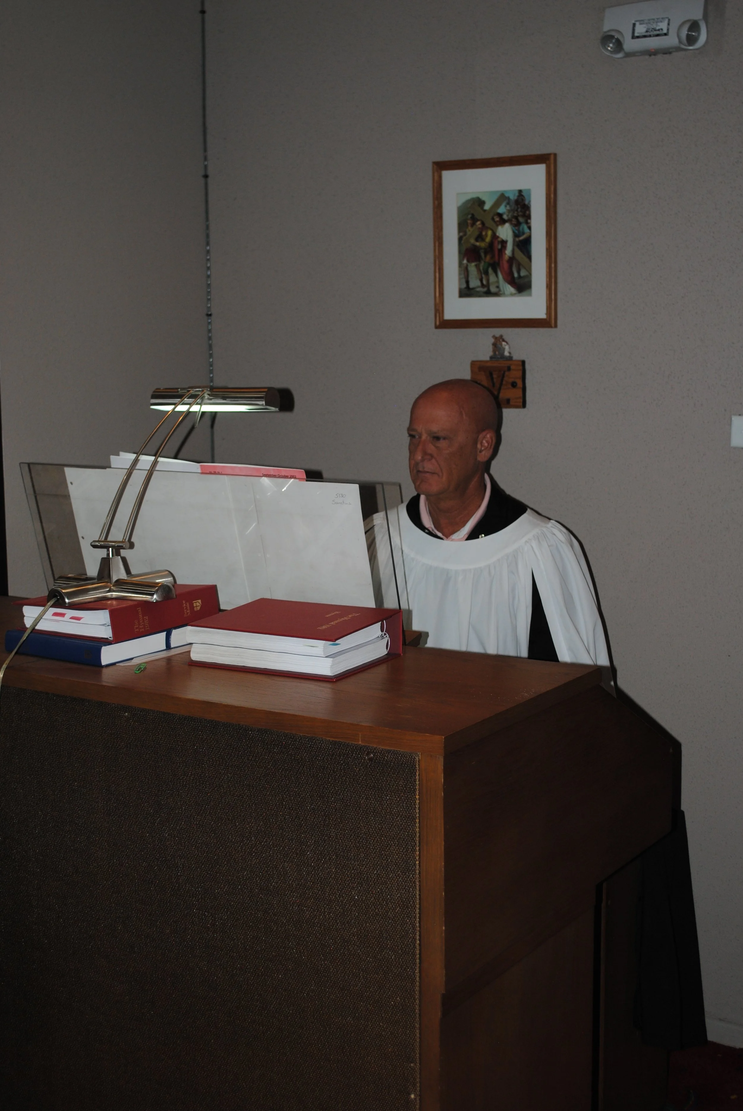 A man in choir robes sitting at a table with books and papers, in a room with religious artwork and a framed picture on the wall.