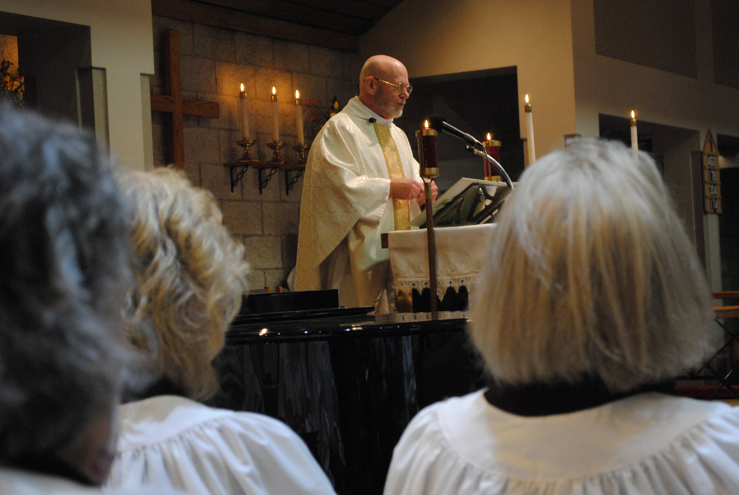 A priest standing at an altar inside a church, with hands folded, during a religious service. There are lit candles and a cross on the wall behind him. The photo is taken from the perspective of the congregation, with the heads of several attendees i