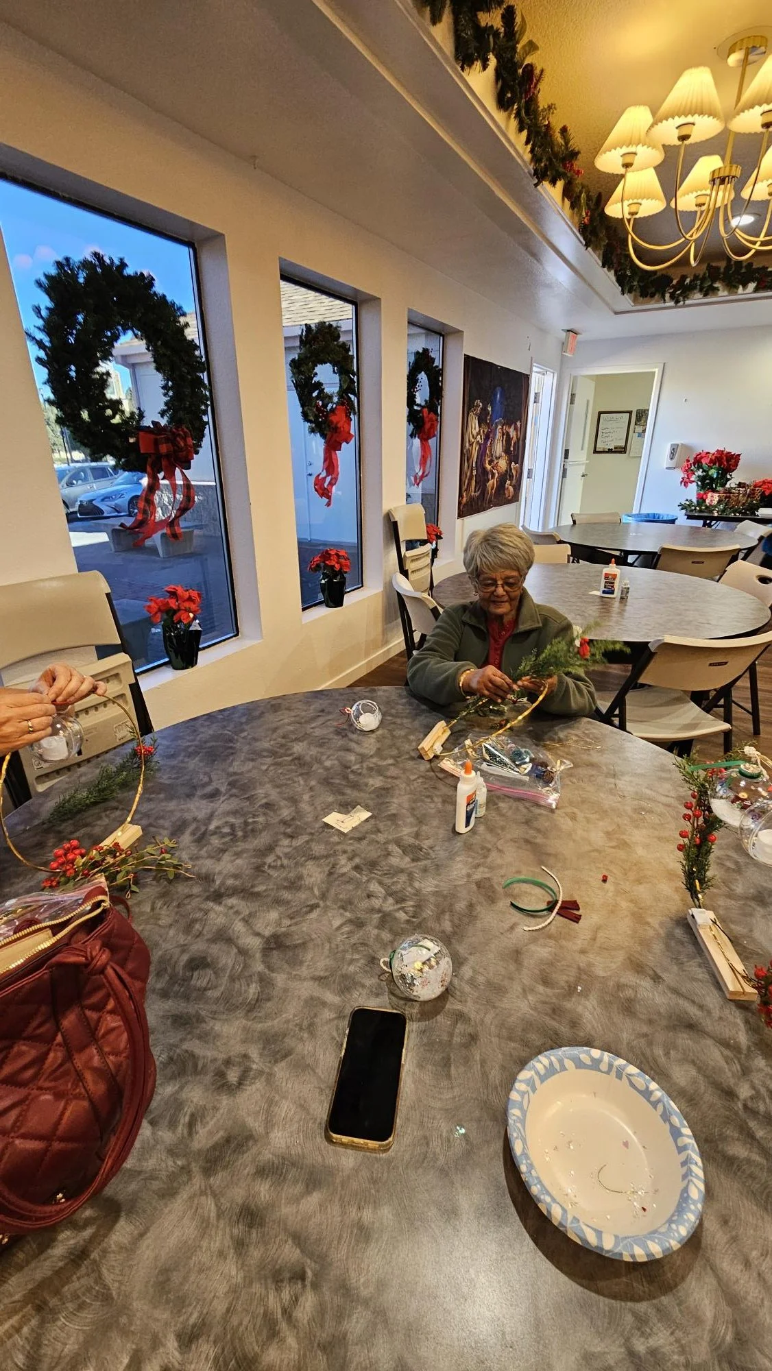 A cozy holiday scene with an elderly woman decorating a wreath at a round table inside a well-lit room. The room is decorated with Christmas wreaths on the windows and garland on the ceiling. There are poinsettia plants and Christmas theme decoration