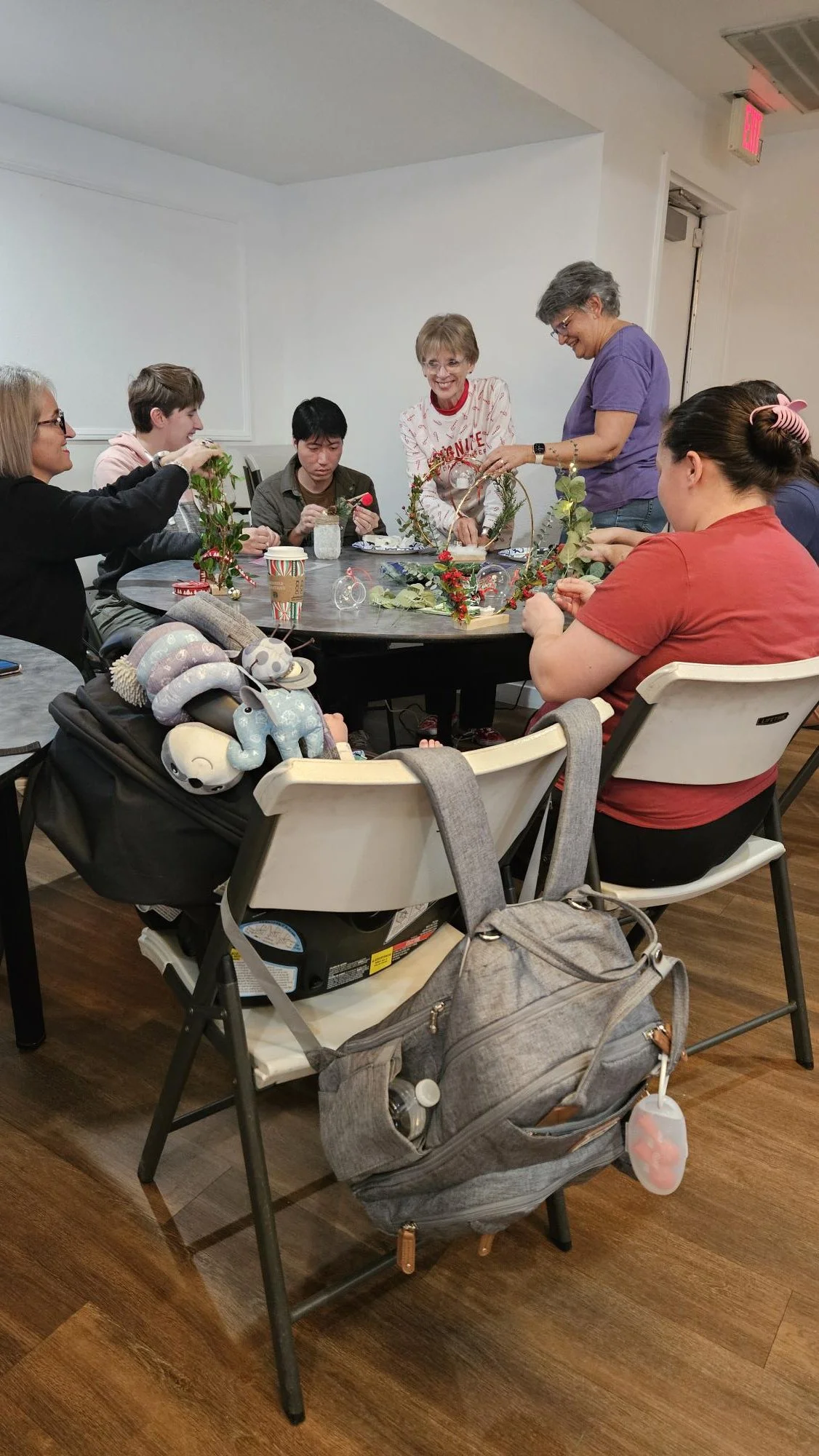 A group of people sitting around a table decorating a Christmas wreath with holiday ornaments and greenery, in a room with hardwood floors and white walls.