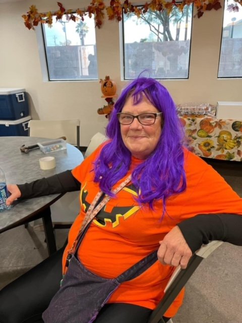 A woman with purple hair and glasses, dressed in an orange Halloween shirt with a jack-o'-lantern face, sitting at a table in a decorated room.