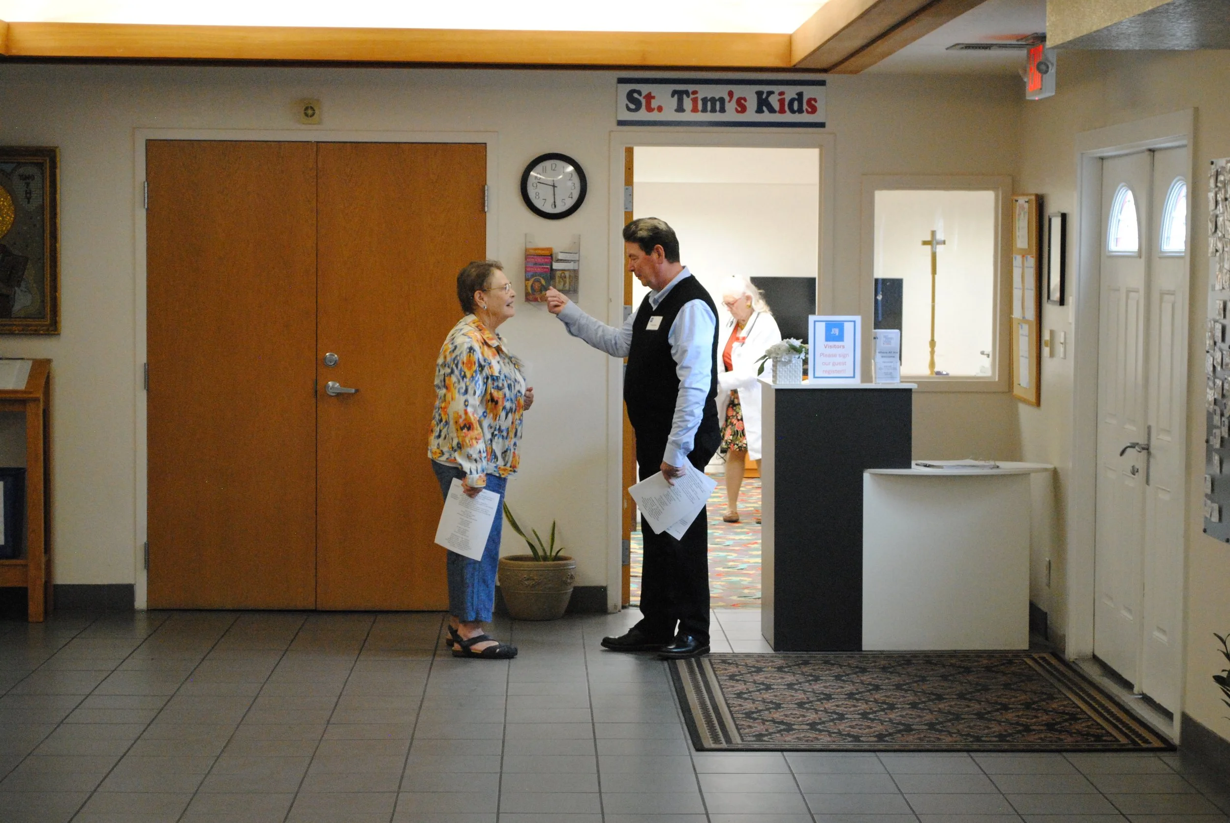 A woman and a man having a conversation at an indoor location, standing near a doorway labeled 'St. Tim's Kids,' with a clock on the wall and a woman in the background.
