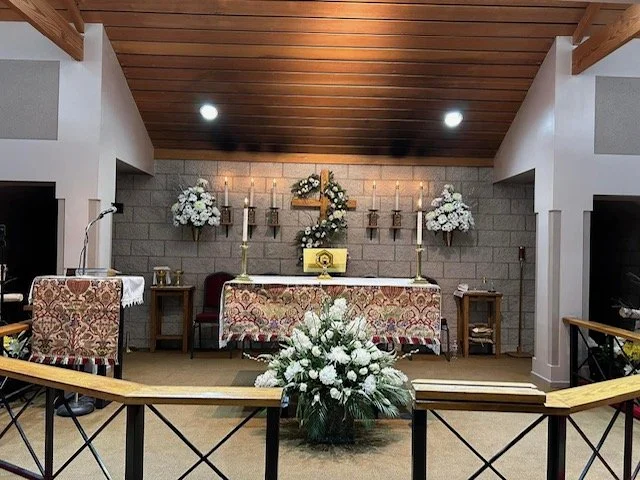 Interior of a chapel with an altar decorated with flowers and candles, set against a brick wall with religious symbols, and a wooden ceiling.
