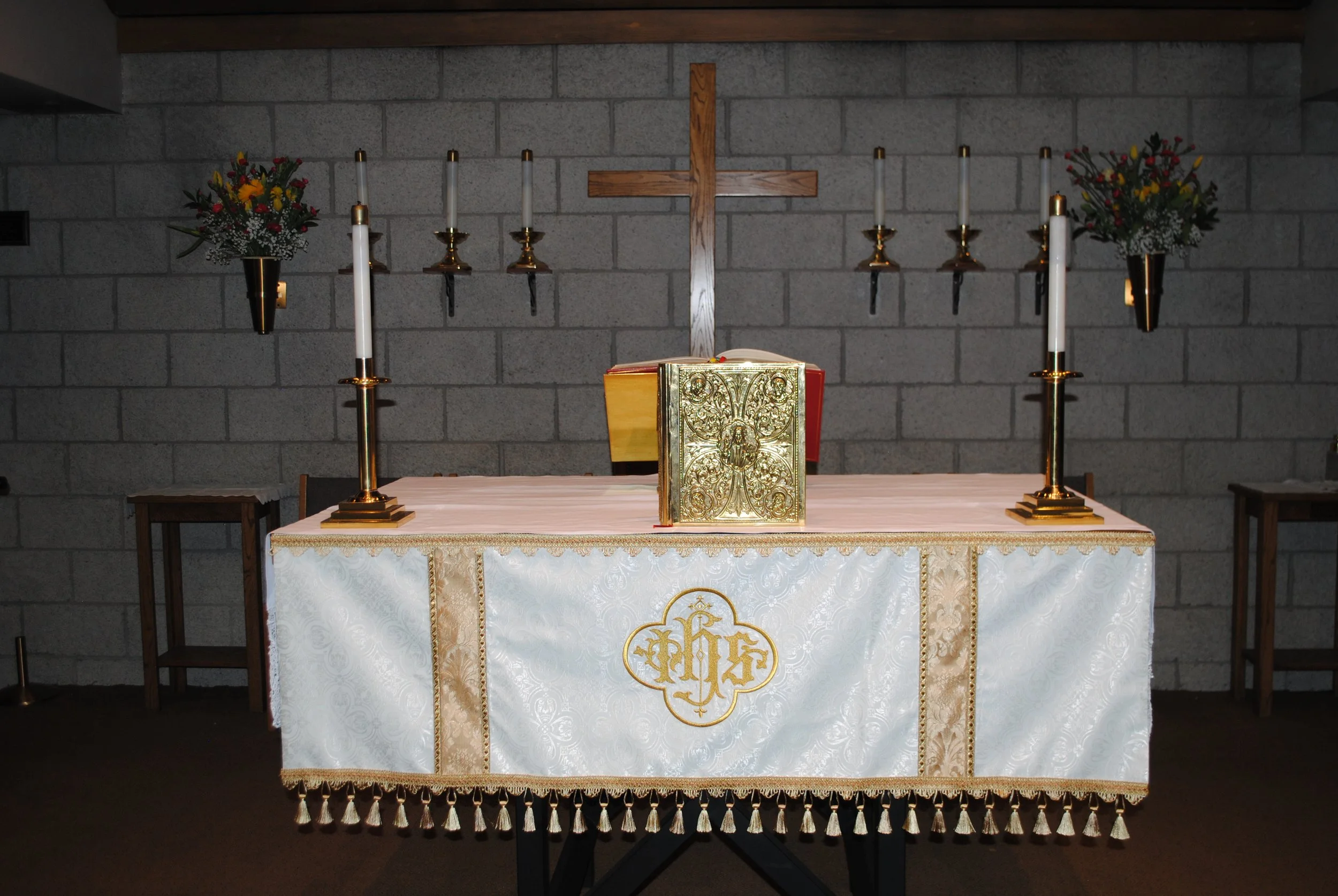 An altar with a wooden cross on a cinder block wall, decorated with white and gold cloth, a gold book, and surrounded by six tall white candles with gold accents, and two bouquets of colorful flowers in black vases.