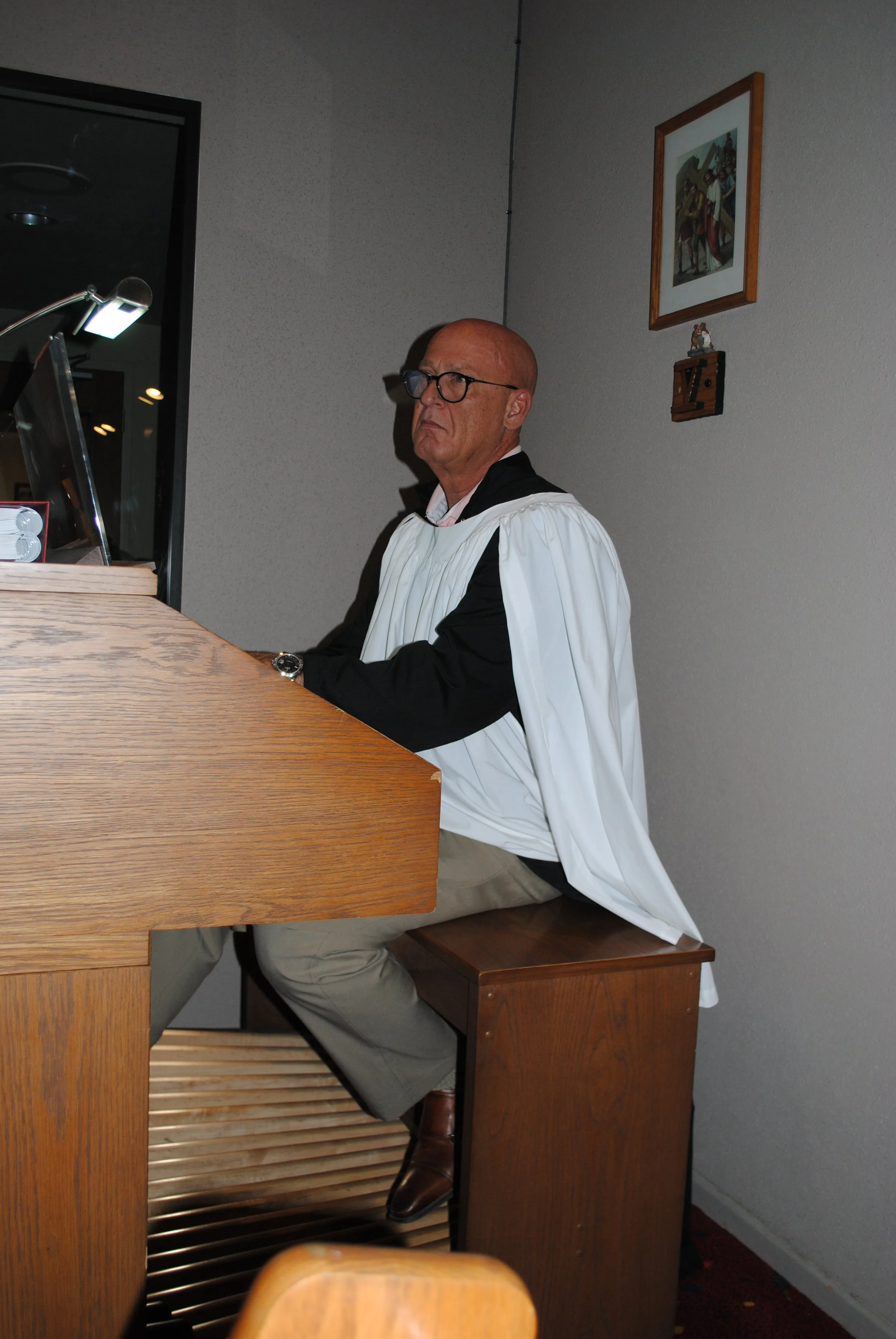 A man with glasses, dressed in a ceremonial robe, sitting at a wooden table in a room with gray walls, a framed picture on the wall, and a wooden bench.