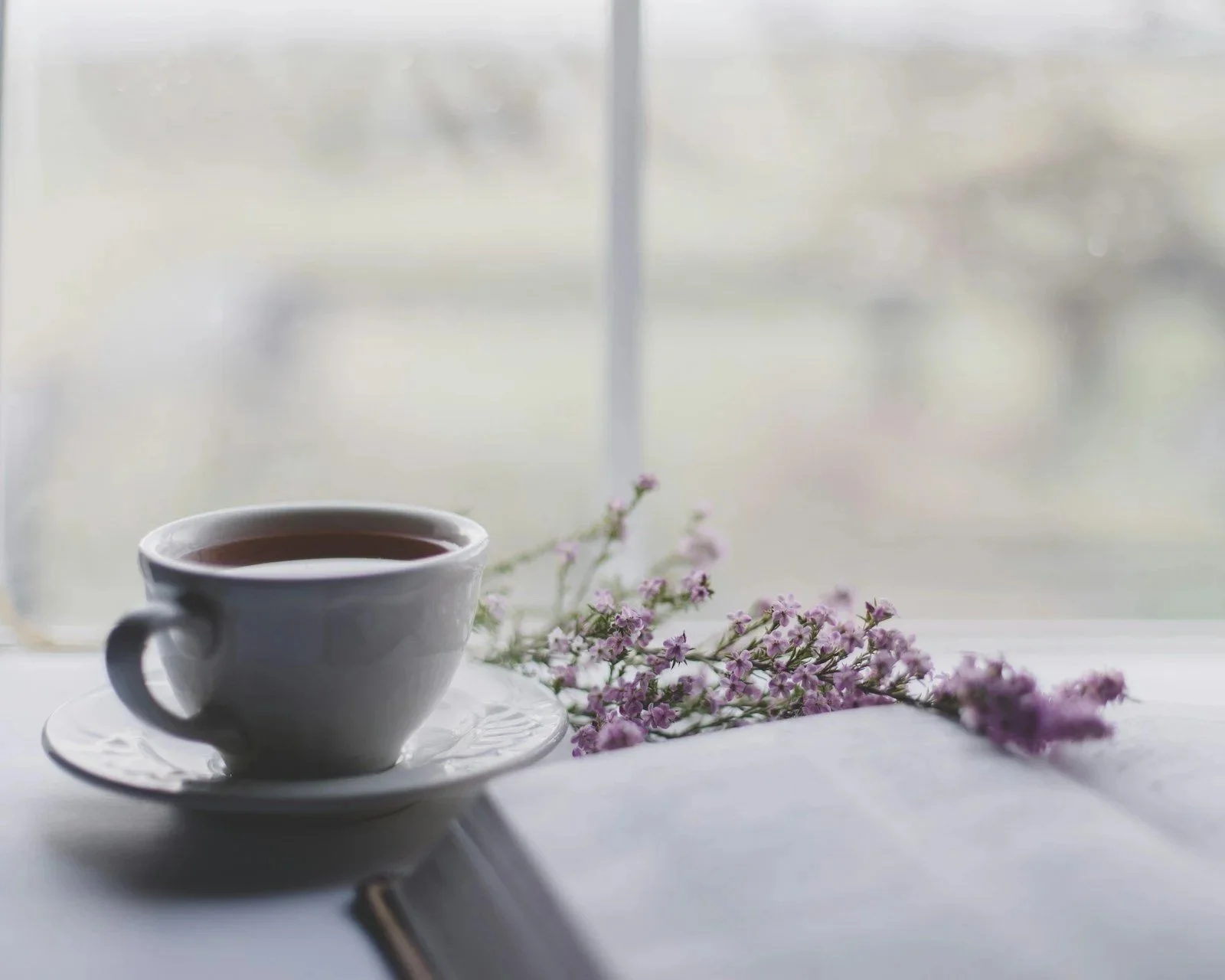 A white cup filled with tea on a saucer, placed on a windowsill with purple flowers beside it, with a blurred outdoor scene in the background.