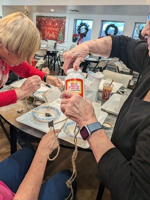 Two women are making holiday-themed crafts at a table, using glue and string, with festive decorations and a "Merry Christmas" sign in the background.