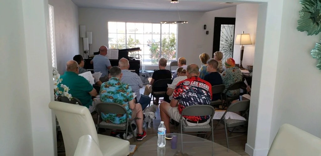 Group of people sitting in a cozy room, attending a music or singing session with a pianist at the front near large window, some reading sheet music.