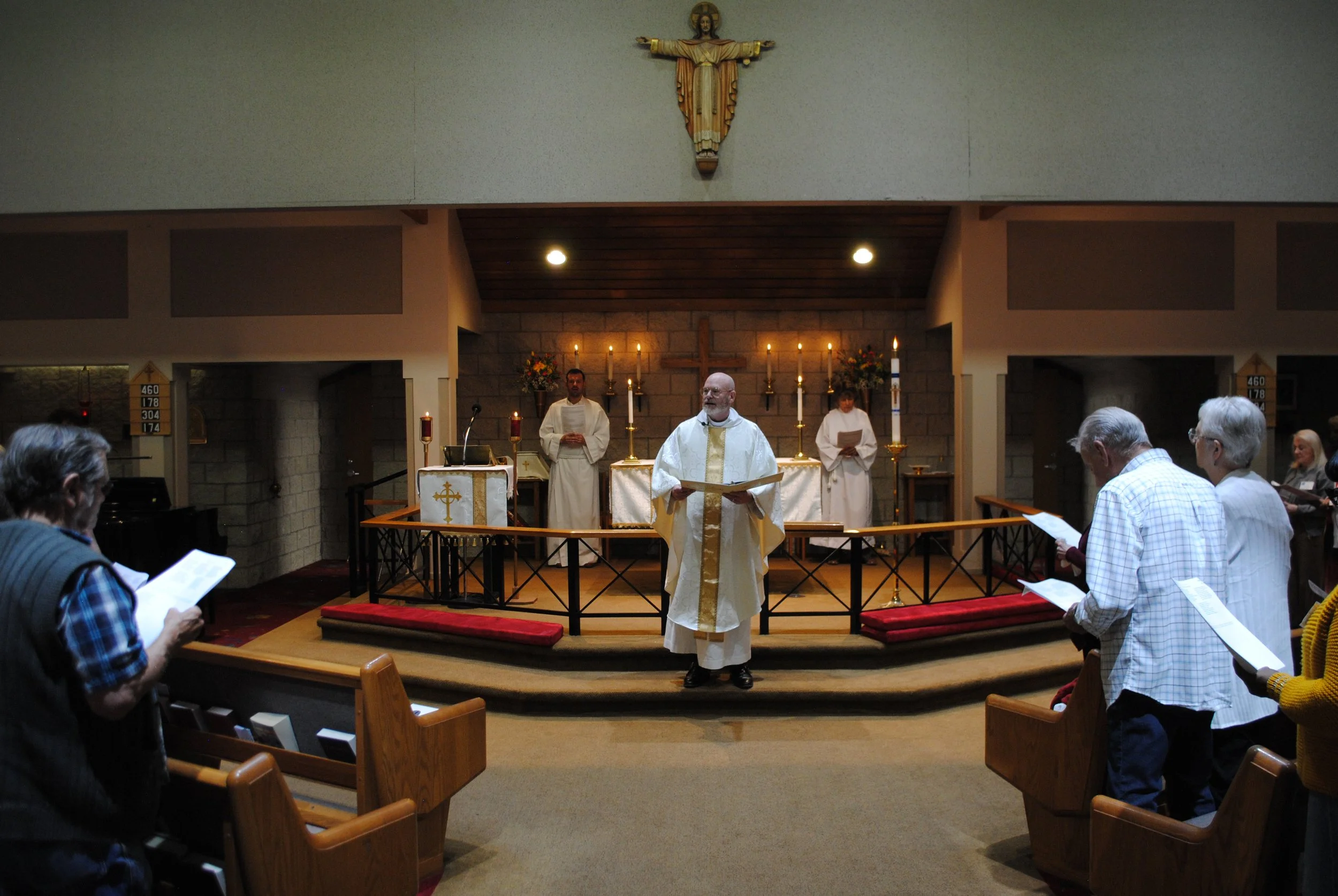 A congregation inside a church during a religious service, with a priest in white robes standing in front of an altar decorated with candles and flowers, and other clergy members behind him. People are holding hymnals or programs, facing the altar.