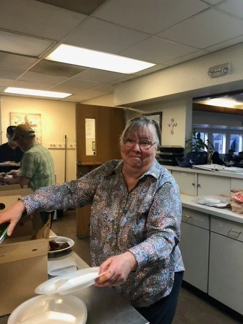 A woman with glasses and a floral blouse stacking disposable plates in a cafeteria or cafeteria-style setting.