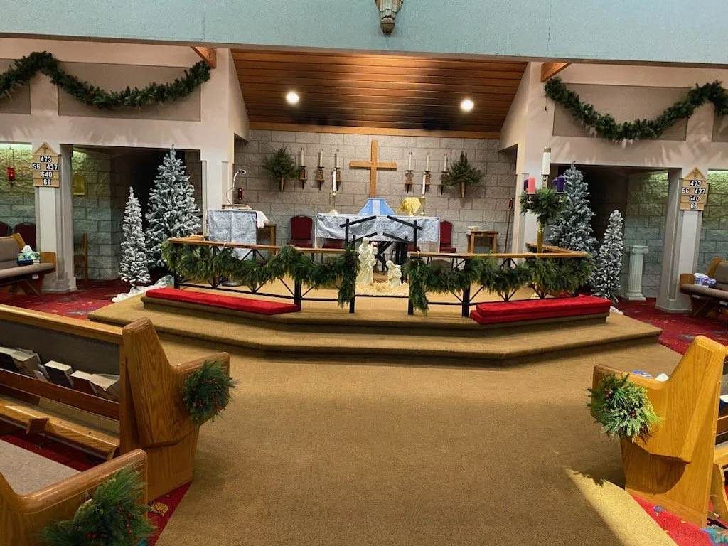 Decorated church altar with Christmas decorations, including miniature snow-covered trees, garlands, and candles, with a wooden cross on the wall behind.