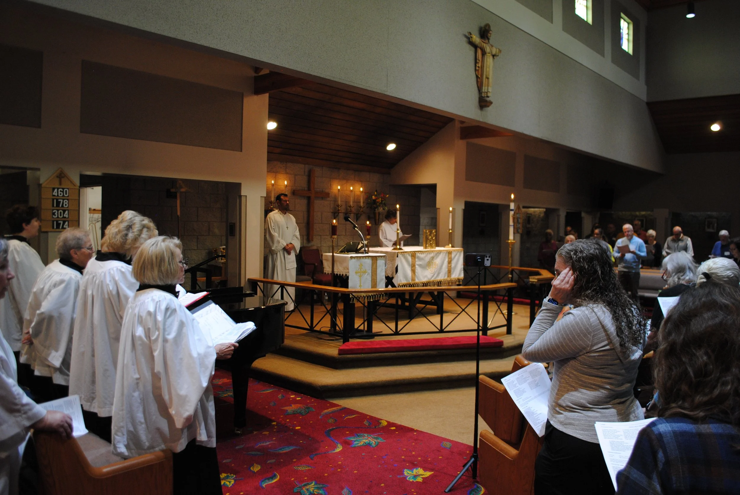 Inside a church during a religious service, with a congregation of people, some wearing choir robes, facing an altar with candles, a cross, and religious icons. The priest and an altar server are present, with one reading from a book.