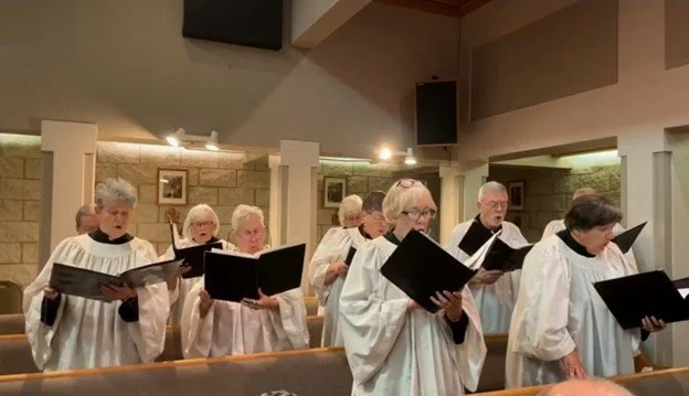 A group of senior choir singers in white robes singing in a church.