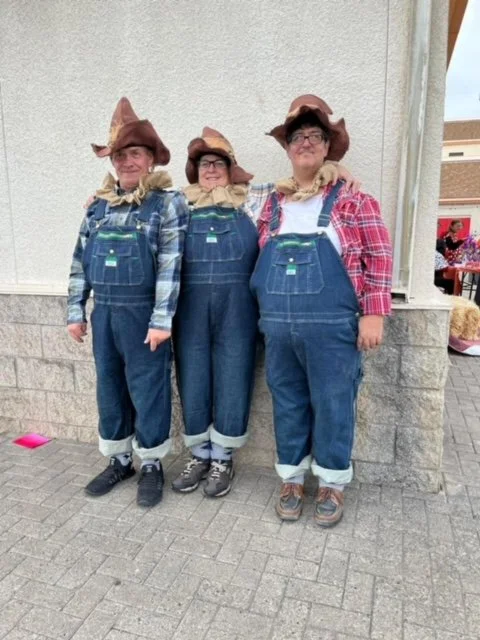 Three people dressed in overalls and cowboy hats standing outdoors against a building wall.